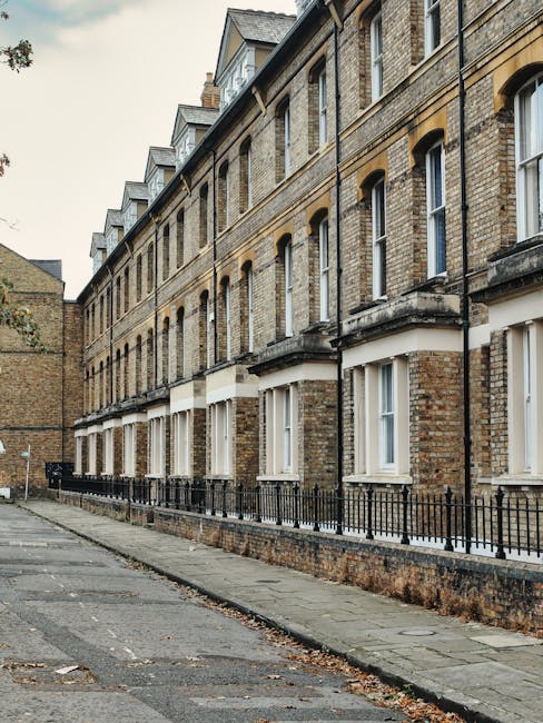 This image depicts a row of traditional brick residential buildings along a narrow sidewalk in Kentish Town. The buildings feature multiple stories with large, white-framed sash windows, some with window sills made of stone or concrete. The facades are constructed from yellowish-brown bricks, with decorative molding and arched window openings on the upper floors. The sidewalk is paved with concrete slabs and bordered by a low black metal railing, separating it from the street made of asphalt. The scene appears clean and well-maintained, with no visible dirt or debris. The lighting suggests an overcast day, casting soft, diffused light over the scene. The image is relevant for content related to domestic or commercial cleaning, exemplifying typical residential exteriors in Kentish Town. Kentish Town Cleaners offers surface cleaning, deep cleaning, and sanitisation services for such properties to maintain hygiene and appearance.