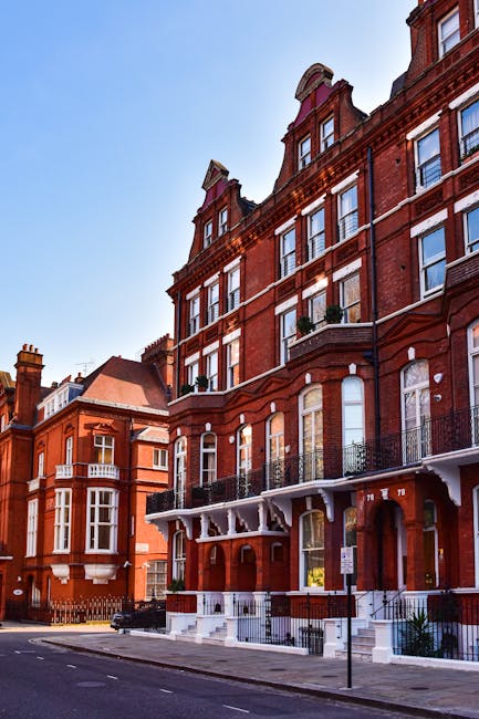 Photograph of a multi-story red brick building with large windows, decorative white trim, and black wrought-iron balconies along the façade, situated on a quiet street under a clear blue sky. The building's exterior appears well-maintained, with clean surfaces and no visible dirt or damage. The lower steps and entrance area are painted white, contrasting with the brickwork. Nearby similar historic buildings are visible, contributing to an urban residential setting. This image exemplifies the type of property that might benefit from professional domestic cleaning services such as end of tenancy cleaning provided by Kentish Town Cleaners, highlighting clean and well-kept surfaces suitable for deep cleaning or sanitisation processes.
