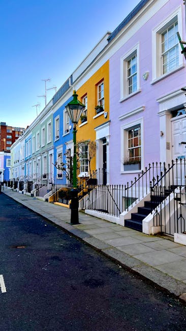 This image depicts a row of traditional brick residential buildings along a narrow sidewalk in Kentish Town. The buildings feature multiple stories with large, white-framed sash windows, some with window sills made of stone or concrete. The facades are constructed from yellowish-brown bricks, with decorative molding and arched window openings on the upper floors. The sidewalk is paved with concrete slabs and bordered by a low black metal railing, separating it from the street made of asphalt. The scene appears clean and well-maintained, with no visible dirt or debris. The lighting suggests an overcast day, casting soft, diffused light over the scene. The image is relevant for content related to domestic or commercial cleaning, exemplifying typical residential exteriors in Kentish Town. Kentish Town Cleaners offers surface cleaning, deep cleaning, and sanitisation services for such properties to maintain hygiene and appearance.