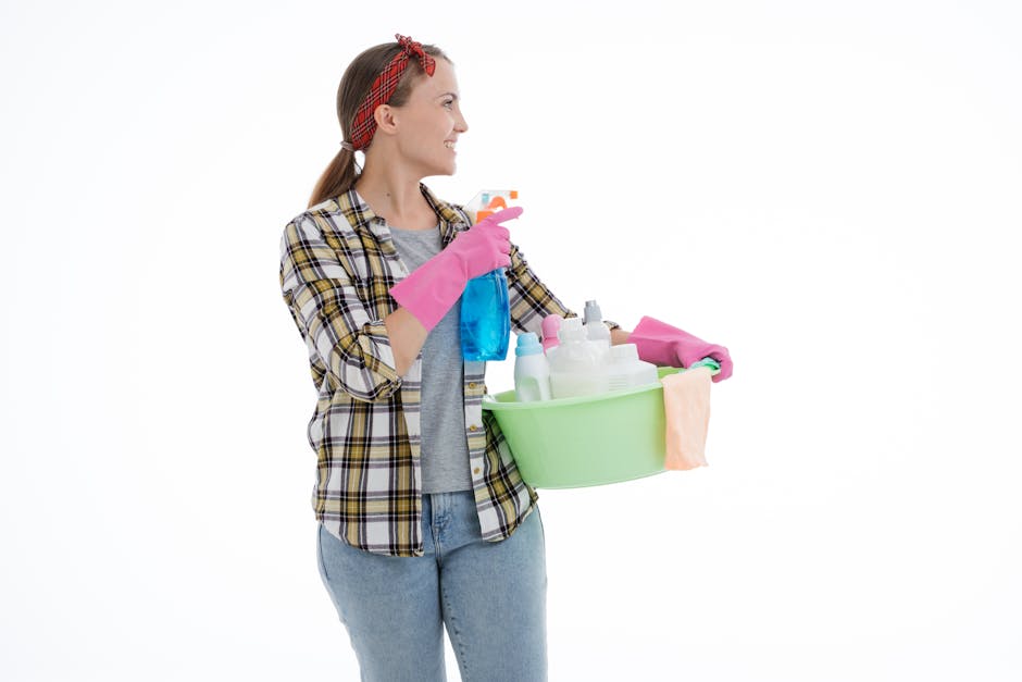 A woman with light skin and brown hair tied back with a red headband, dressed in a plaid shirt over a grey top and light blue jeans, is holding a green plastic basin filled with various cleaning supplies including spray bottles, cloths, and sponges. She is wearing pink rubber gloves and is smiling while looking to her left. The background is plain white, emphasizing her preparation for domestic cleaning tasks, which aligns with surface cleaning and sanitisation processes promoted by Kentish Town Cleaners, relevant to avoiding hidden charges in Kentish Town cleaning quotes.