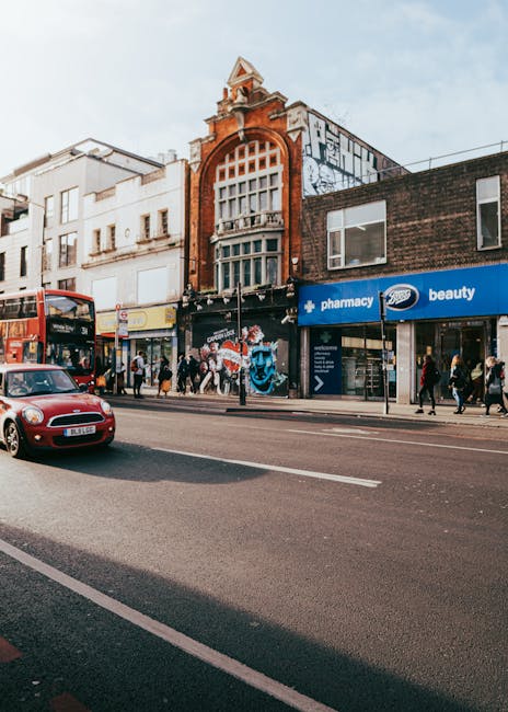 Exterior view of a busy commercial street in Kentish Town, showing a mix of buildings with decorative brickwork and large windows. The storefronts include a pharmacy and a beauty shop, both with modern signage. The sidewalk is populated with pedestrians, some entering or exiting shops, and others walking along the street. A red double-decker bus and a small red car are traveling on the road in front of the buildings. The scene is lit by natural daylight, highlighting the clean and well-maintained surfaces of the shop windows, pavement, and road. The overall atmosphere suggests a vibrant, urban environment with active foot and vehicle traffic, and the image emphasizes the importance of regular cleaning and maintenance as part of maintaining a hygienic and visually appealing commercial area, as promoted by Kentish Town Cleaners in relation to Camden Council rules for bulk waste removal.