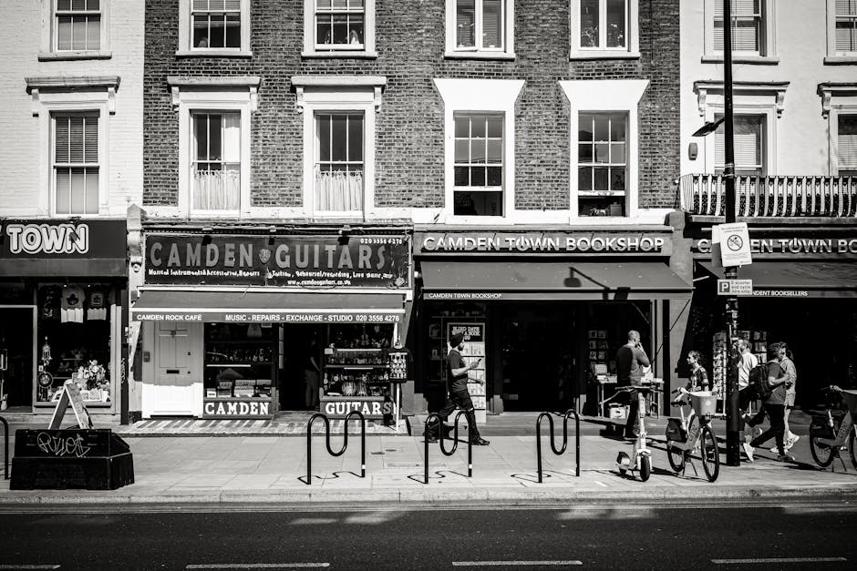 A black and white street view of commercial storefronts in Kentish Town, featuring signage for a guitar shop named Camden Guitars and a bookstore called Camden Town Bookshop. The pavement displays several pedestrians walking past, some carrying shopping bags or backpacks, and two bicycles secured to bike racks in front of the shops. The buildings above the shops have classical brick facades with evenly spaced windows, some with white window frames and blinds. The shopfronts are adorned with awnings, with the guitar shop's window showcasing various musical instruments and accessories. The street scene is well-lit, highlighting the clean, orderly sidewalk surfaces and the organized arrangement of street furniture, such as bike racks and a parking meter. This image emphasizes the importance of surface cleaning and maintenance in urban environments, aligning with the themes of domestic and commercial cleaning services provided by Kentish Town Cleaners, especially in accordance with Camden Council rules for bulk waste removal.