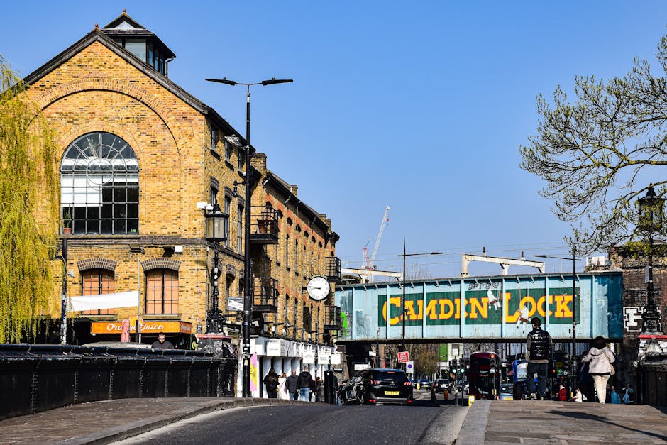 Exterior view of a busy commercial street in Kentish Town, showing a mix of buildings with decorative brickwork and large windows. The storefronts include a pharmacy and a beauty shop, both with modern signage. The sidewalk is populated with pedestrians, some entering or exiting shops, and others walking along the street. A red double-decker bus and a small red car are traveling on the road in front of the buildings. The scene is lit by natural daylight, highlighting the clean and well-maintained surfaces of the shop windows, pavement, and road. The overall atmosphere suggests a vibrant, urban environment with active foot and vehicle traffic, and the image emphasizes the importance of regular cleaning and maintenance as part of maintaining a hygienic and visually appealing commercial area, as promoted by Kentish Town Cleaners in relation to Camden Council rules for bulk waste removal.