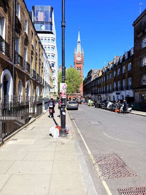 A city street scene in Kentish Town, featuring a wide sidewalk with clean, evenly laid concrete slabs and a black metal railing along the edge. There are pedestrians, including a person with a backpac