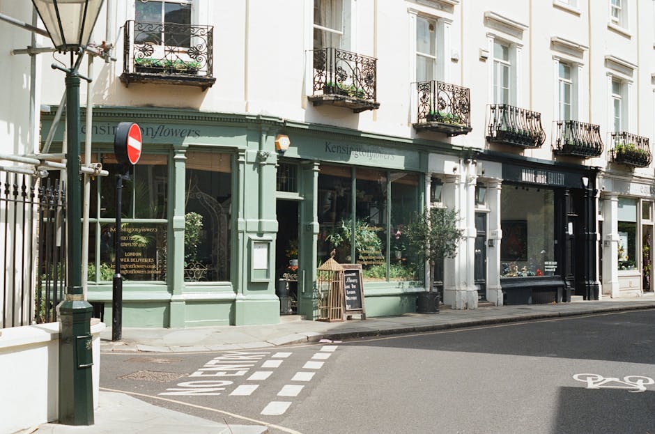Image of a row of retail shops on a city street in daylight, with a flower shop featuring a green storefront, large glass windows displaying plants and flowers, and a blackboard sign outside. The street has a bicycle lane marked with a painted bicycle symbol, and pedestrian crossing lines are visible on the asphalt. The buildings above the shops are white with ornate black iron balconies and large windows, illuminated by natural light. The scene is clean and well-maintained, reflecting a typical urban environment in Kentish Town, with no visible dirt or clutter. Kentish Town Cleaners specializes in surface cleaning and deep cleaning services to address common accessibility problems for flats and commercial spaces.