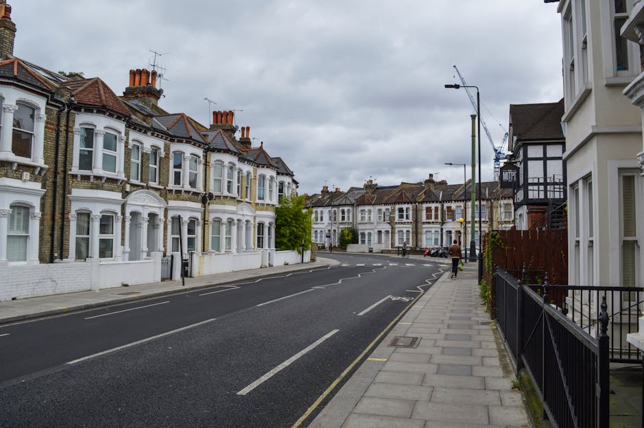 A quiet residential street in Kentish Town featuring a row of Victorian-style terraced houses with white and brick facades, bay windows, and red tiled roofs. The street is paved with dark asphalt and has clearly marked parking bays along the curb. The sidewalk is made of concrete slabs with a black metal railing separating it from the road. Overcast sky with grey clouds provides diffused lighting, highlighting the clean, well-maintained surfaces of the buildings and pavement. Visible features include street lamps, a few pedestrians, and some parked bicycles, exemplifying typical urban domestic surroundings where cleaning and maintenance are essential for hygiene and appearance, as managed by Kentish Town Cleaners.