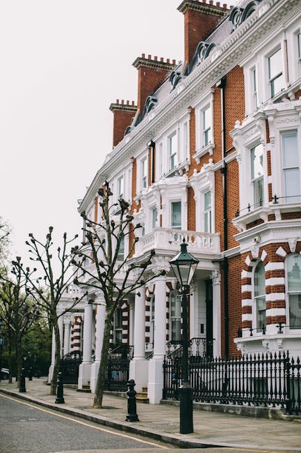A row of Victorian-style residential buildings along Crowndale Road in Kentish Town, featuring red brick facades with white decorative architectural elements such as window frames, cornices, and balustrades. The buildings have tall, arched windows and ornate details, with a black wrought iron fence running along the sidewalk. Leafless trees line the street, and a vintage-style street lamp stands near the entrance of one property. The scene is captured during daylight with overcast sky, and the image emphasizes exterior surfaces that are well-maintained and clean, representing the kind of property that Kentish Town Cleaners might service for estate cleaning and maintenance. The overall setting highlights the historic charm and meticulous exterior presentation typical of this area.