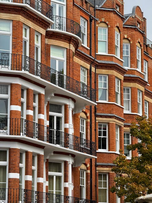 Exterior view of a multi-storey residential building with red brick facade, white accents, and black wrought-iron balconies. The building features large rectangular windows with white frames, some with partially drawn curtains, and a curved balcony on each floor supported by white columns. The scene is well-lit with natural daylight, and a leafy tree with green and yellow leaves is visible in the lower right corner. The image conveys the tidy and maintained appearance of the building's exterior. Kentish Town Cleaners offers estate cleaning services for such properties, ensuring surfaces remain clean and well-kept.