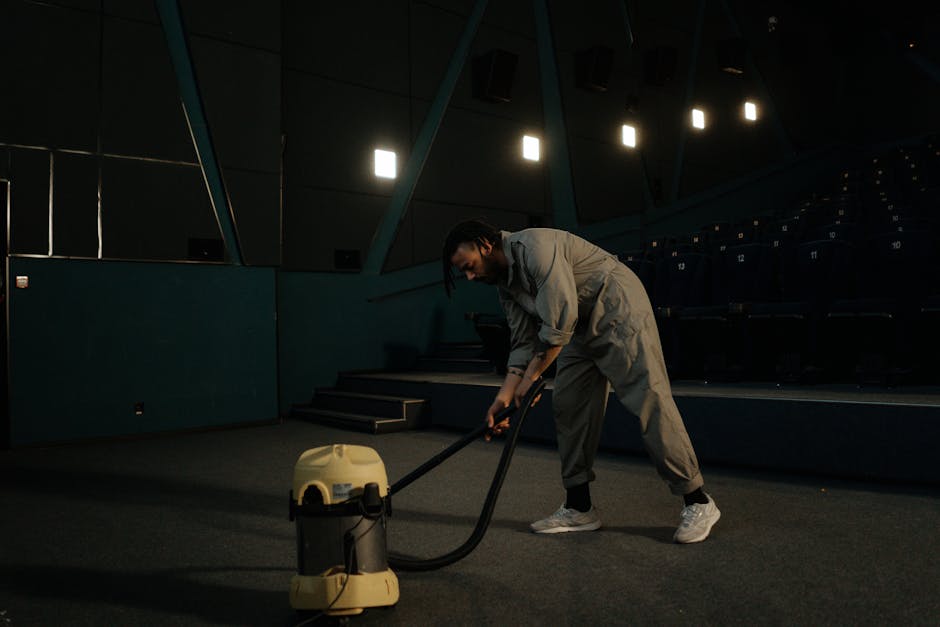 A person wearing a light grey jumpsuit and white sneakers is performing deep cleaning or sanitisation in an auditorium or lecture hall. They are using a portable vacuum or extraction device to clean the carpeted floor near the stairs, with their focus directed at the surface. The auditorium features dark blue theatre seats arranged in rows, with numbered seat markers visible on the right side. The walls are covered with panels, and the room is dimly lit by overhead lights, creating a professional and clean environment. The scene emphasizes surface cleaning and maintenance by Kentish Town Cleaners, demonstrating thorough hygiene procedures in a commercial setting.