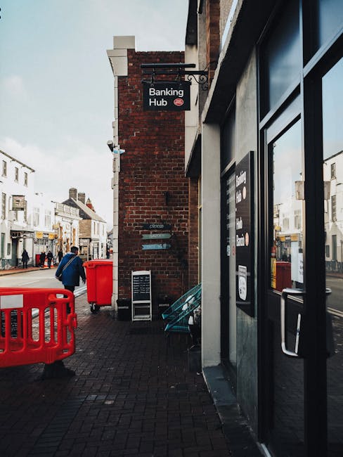 A person wearing a light grey jumpsuit and white sneakers is performing deep cleaning or sanitisation in an auditorium or lecture hall. They are using a portable vacuum or extraction device to clean the carpeted floor near the stairs, with their focus directed at the surface. The auditorium features dark blue theatre seats arranged in rows, with numbered seat markers visible on the right side. The walls are covered with panels, and the room is dimly lit by overhead lights, creating a professional and clean environment. The scene emphasizes surface cleaning and maintenance by Kentish Town Cleaners, demonstrating thorough hygiene procedures in a commercial setting.