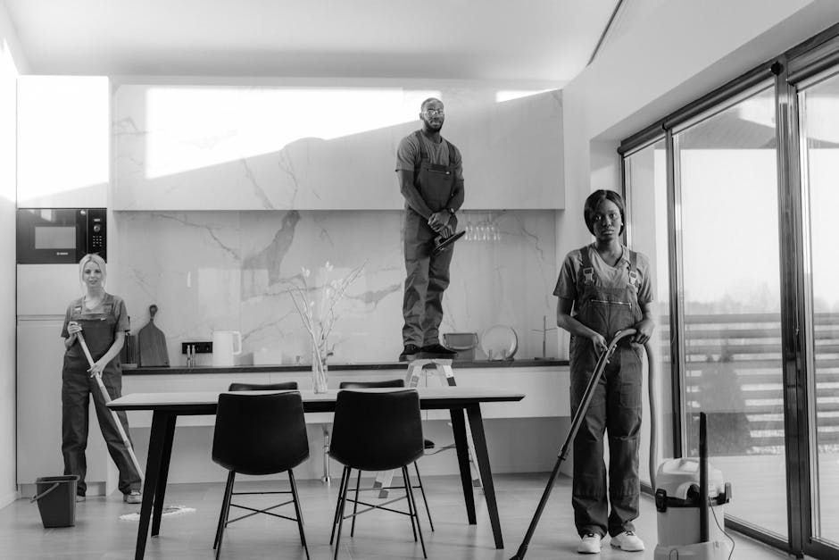 A black-and-white photograph of three professional cleaners in an open-plan kitchen and dining area. The room features a modern design with a large marble backsplash and countertop, and glass sliding doors leading outside. One cleaner, standing on a step ladder, is wiping the upper kitchen cabinets. Another is holding a mop with a bucket nearby, focused on cleaning the wooden or tile floor. The third cleaner, standing at the side, holds a cloth or mop handle, possibly scrubbing the floor. The space is well-lit with natural light coming through the large glass doors. Visible cleaning tools include mop, bucket, cloths, and spray bottles. The professional cleaners are dressed in uniforms, demonstrating thorough surface cleaning and hygiene practices. This image illustrates a deep cleaning or sanitisation process within a domestic setting, relevant to legal end-of-tenancy cleaning obligations, with a focus on surface cleaning, sanitation, and maintaining high hygiene standards. Kentish Town Cleaners' name is naturally integrated into this scene, showcasing their commitment to professional cleaning services.