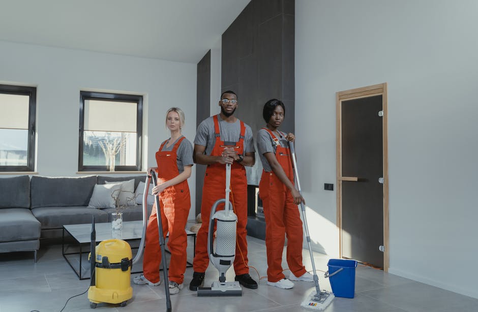 A black-and-white photograph of three professional cleaners in an open-plan kitchen and dining area. The room features a modern design with a large marble backsplash and countertop, and glass sliding doors leading outside. One cleaner, standing on a step ladder, is wiping the upper kitchen cabinets. Another is holding a mop with a bucket nearby, focused on cleaning the wooden or tile floor. The third cleaner, standing at the side, holds a cloth or mop handle, possibly scrubbing the floor. The space is well-lit with natural light coming through the large glass doors. Visible cleaning tools include mop, bucket, cloths, and spray bottles. The professional cleaners are dressed in uniforms, demonstrating thorough surface cleaning and hygiene practices. This image illustrates a deep cleaning or sanitisation process within a domestic setting, relevant to legal end-of-tenancy cleaning obligations, with a focus on surface cleaning, sanitation, and maintaining high hygiene standards. Kentish Town Cleaners' name is naturally integrated into this scene, showcasing their commitment to professional cleaning services.