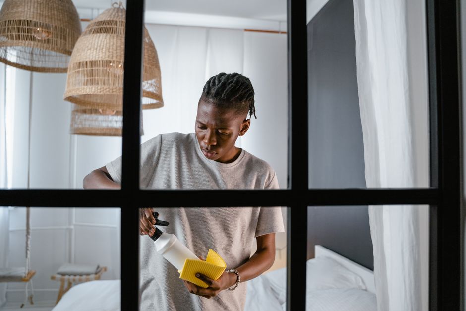 A person dressed in a grey shirt and orange overalls is using a yellow vacuum cleaner with a grey hose to clean a living room. The room features a large grey fabric sofa with white and grey cushions, 