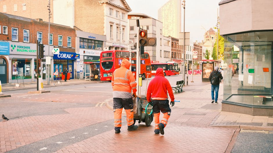Two street cleaning workers dressed in orange high-visibility uniforms are pushing a cart with cleaning tools along a city sidewalk. The scene shows a busy urban area in Kentish Town with a red double-decker bus, shops, pedestrians, and traffic lights visible in the background. The sidewalk is made of red bricks, and the storefronts include a pharmacy and a building with large glass windows. The lighting suggests early morning or late afternoon. Kentish Town Cleaners emphasizes surface and deep cleaning services, contributing to a clean and hygienic environment aligned with the guidelines for council permits and urban maintenance.