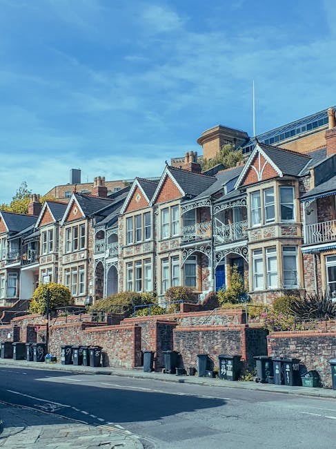 A row of Victorian-style terraced houses in Kentish Town with ornate white detailing on their brick facades, large bay windows, and small front gardens bordered by brick walls. The houses are illuminated by natural daylight against a partly cloudy blue sky. In the foreground, a paved street with a row of black wheelie bins lines the curb, and the overall scene is clean and well-maintained, reflecting the residential area serviced by Kentish Town Cleaners for comprehensive domestic cleaning and surface sanitisation. The image emphasizes the importance of maintaining hygiene standards in historic neighbourhoods, aligning with the knowledge about council permits for cleaning services in the area.