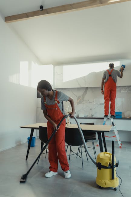 Two professional cleaners wearing red overalls are performing surface cleaning in a modern kitchen with white walls and marble-effect splashbacks. One cleaner is using a vacuum cleaner on the floor, which appears to be a smooth, light-colored laminate surface, while the other is standing on a step ladder, wiping or polishing the high section of the wall or cabinetry with a cloth. The kitchen features a wooden ceiling beam, a large dining table with black chairs, and ample natural light coming from windows. The scene reflects deep cleaning and sanitisation processes, emphasized by the use of quality cleaning tools and the well-maintained, spotless surfaces. This image illustrates the domestic cleaning services offered by Kentish Town Cleaners in Kentish Town as part of urgent same-day cleaning solutions.